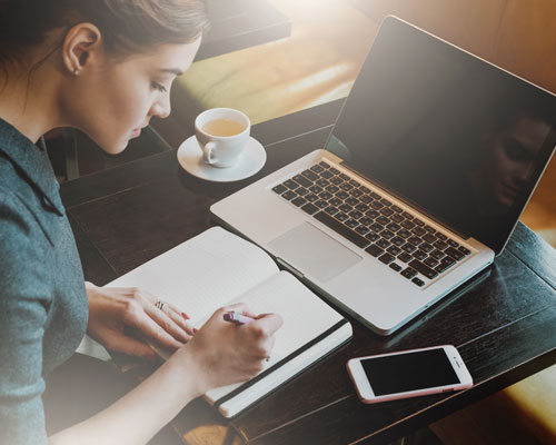 A Woman Writing in Notebook With Laptop on Table